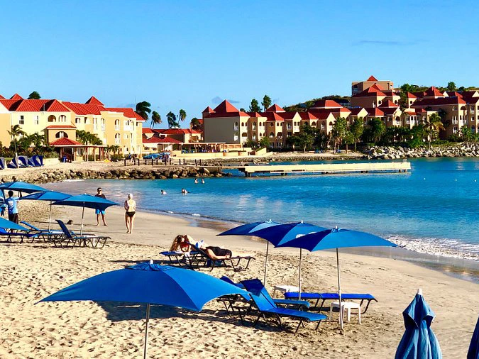 Beachfront view of Divi Little Bay Beach Resort in St. Maarten featuring blue sun umbrellas, lounge chairs, and red-roofed villas along the turquoise shore.