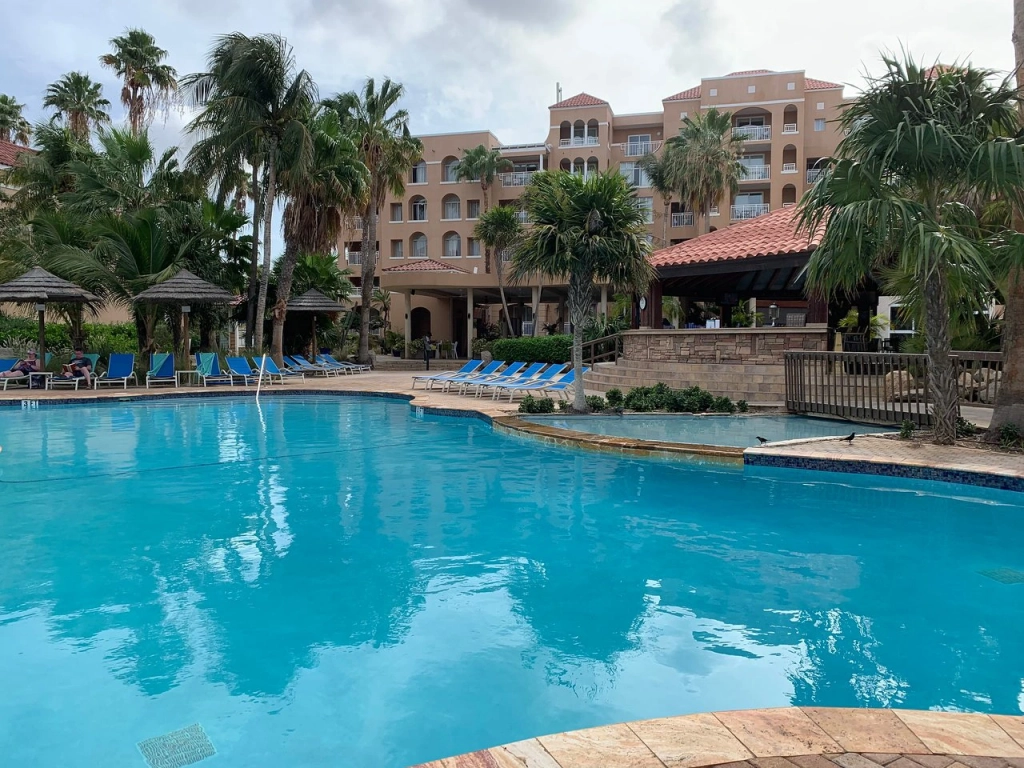 Large outdoor pool area at Divi Village Golf and Beach Resort surrounded by tropical palm trees and blue lounge chairs.