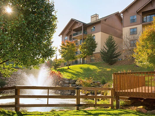 A tranquil pond with a fountain, surrounded by trees and grass, beside a wooden deck at Wyndham Smoky Mountains Resort.