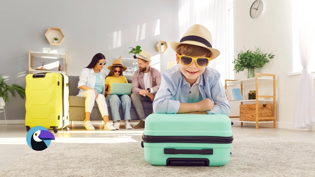 A cheerful boy in sunglasses and a hat lies on a mint green suitcase. In the background, a family sits on a sofa with a yellow suitcase nearby. - Summer Vacation Ideas Feature Image