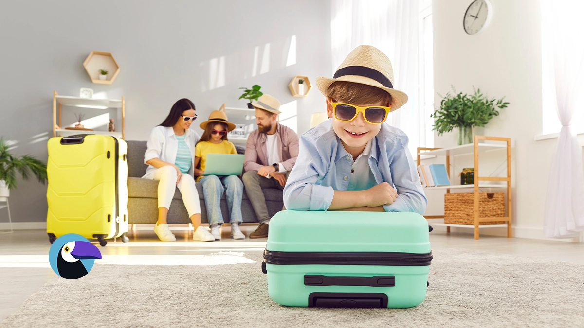 A cheerful boy in sunglasses and a hat lies on a mint green suitcase. In the background, a family sits on a sofa with a yellow suitcase nearby. - Summer Vacation Ideas Feature Image