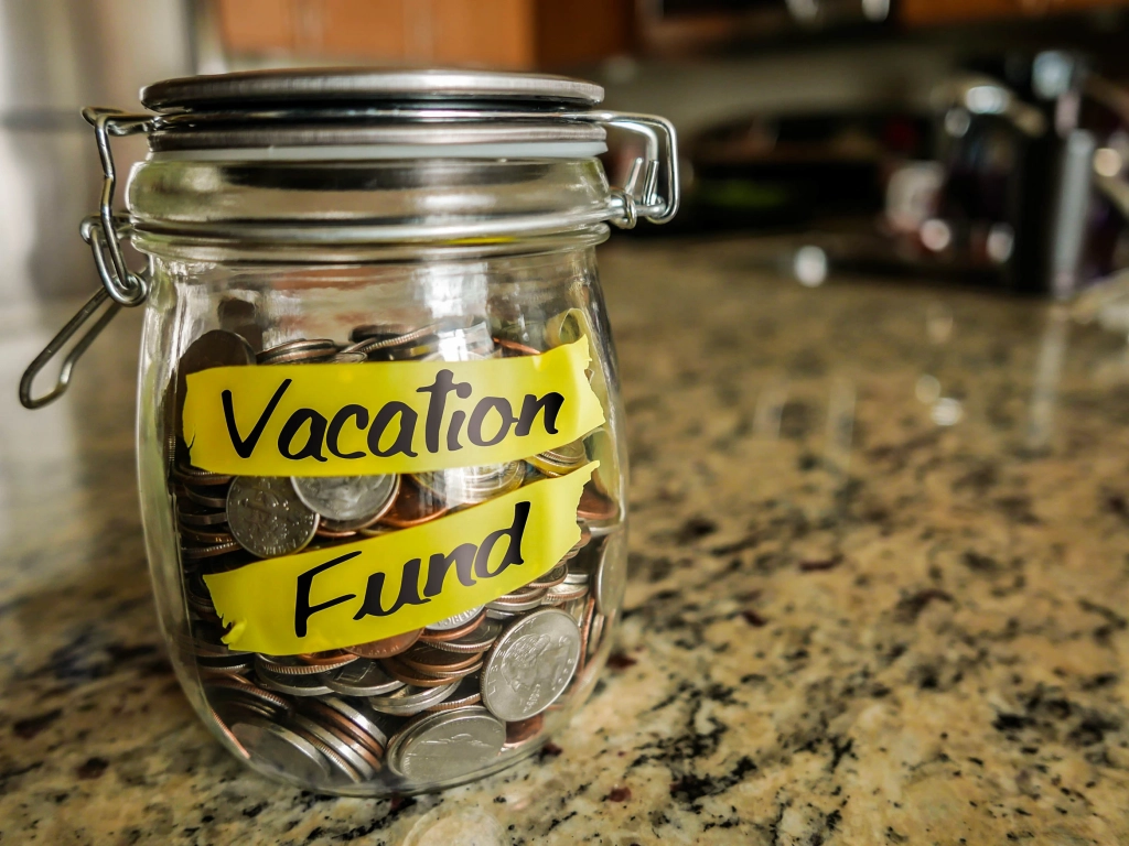 Glass savings jar full of coins with a bright yellow tape label that reads Vacation Fund sitting on a granite countertop, illustrating the concept of timeshare points.