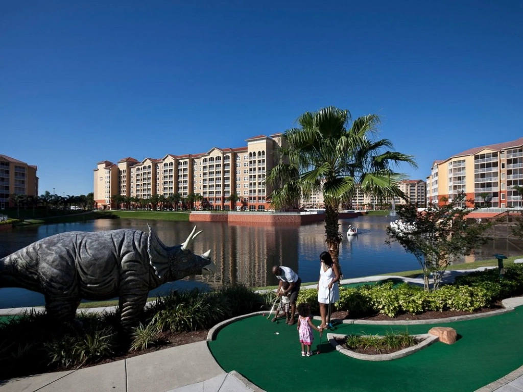 Family playing mini-golf next to a large triceratops dinosaur statue at a lakeside resort