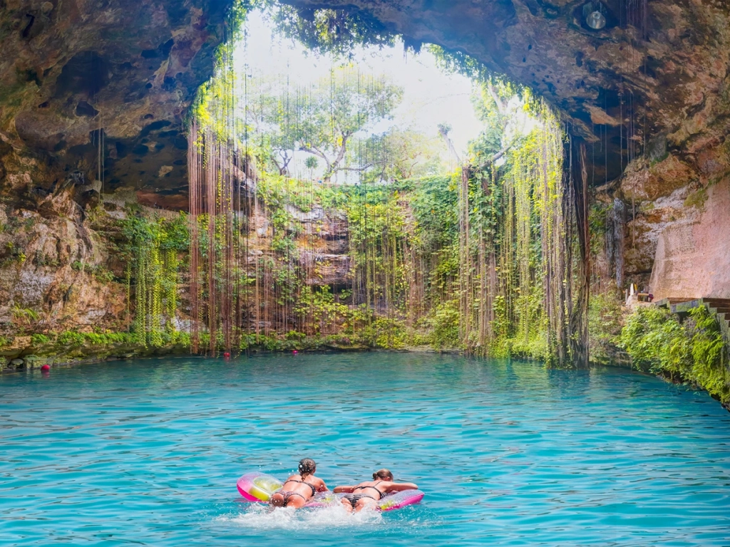 Two people relax on pink floats in a bright blue cenote surrounded by rocky cave walls and hanging vines.