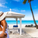 Woman in white dress and hat leading someone toward a white gazebo on a tropical white sand beach with clear blue water.