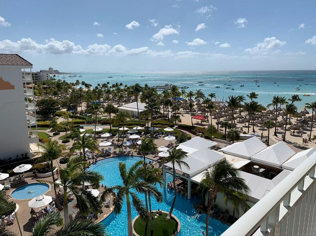 Looking down from a balcony at the marriott aruba resort, you can see a bright blue pool, sandy beach, and calm ocean.