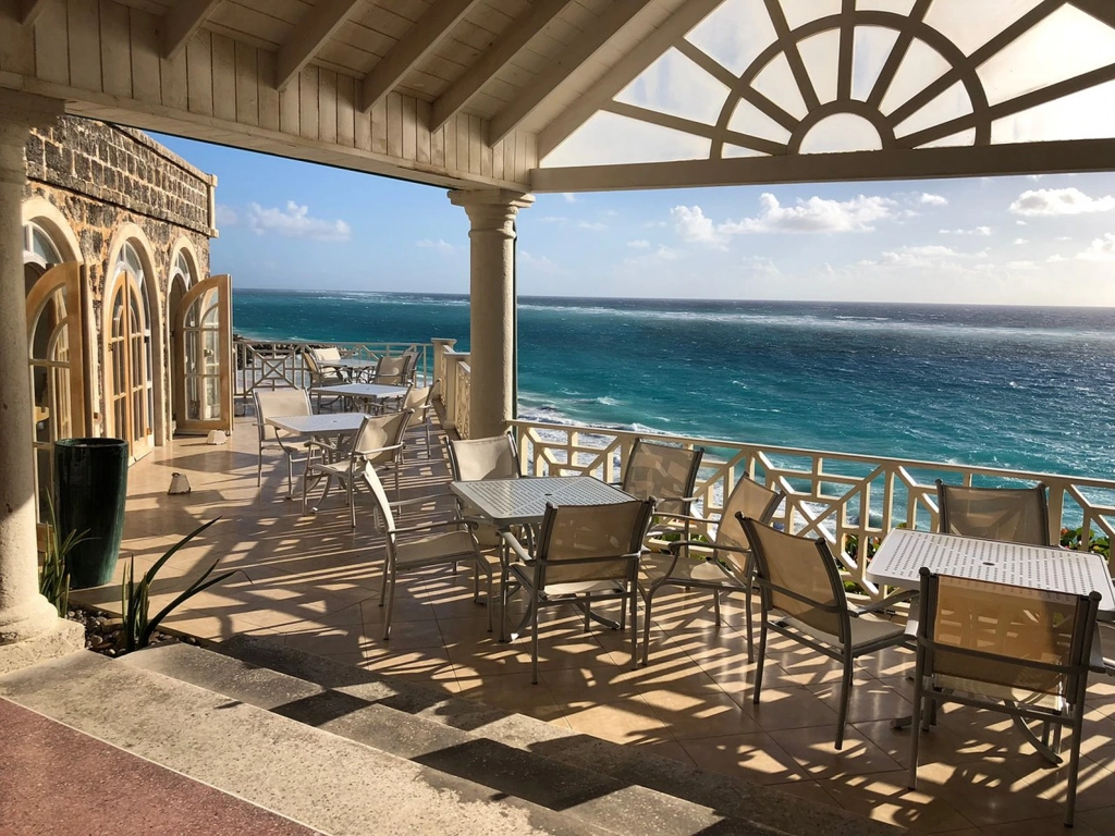 Shaded stone balcony with dining tables overlooking the bright blue ocean at The Crane Resort, a Caribbean timeshare.