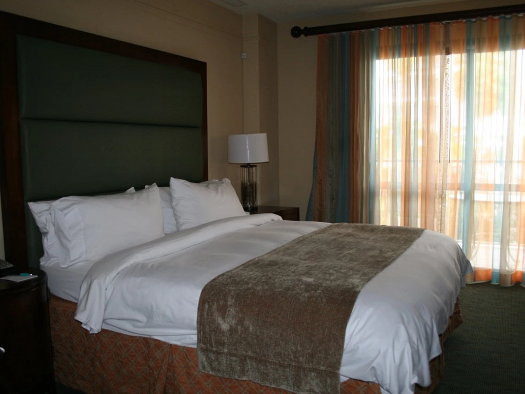 A neatly made hotel bed with a green headboard and sheer striped curtains near the window.