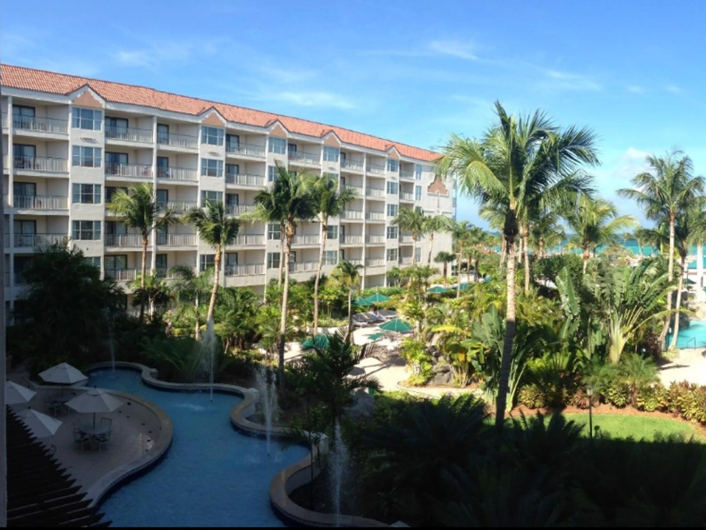 A sunny resort courtyard with a winding blue pool, small water fountains, and tall palm trees in front of a white building.
