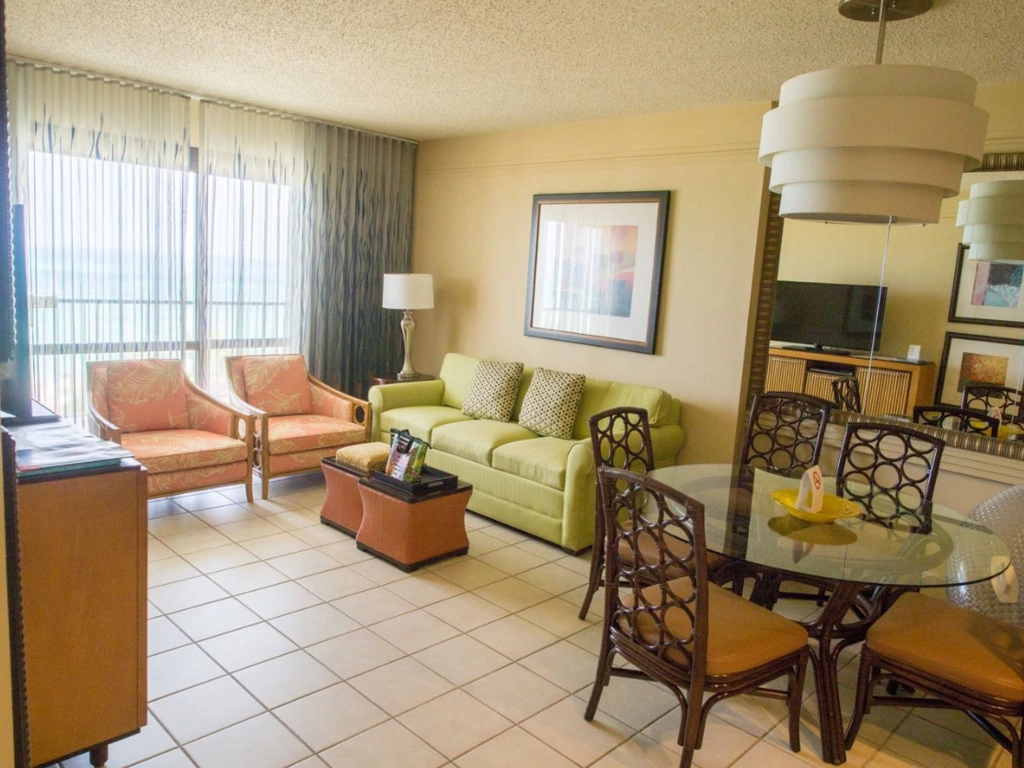 A comfortable hotel living area decorated in tropical greens and oranges, complete with a sofa and glass dining table.