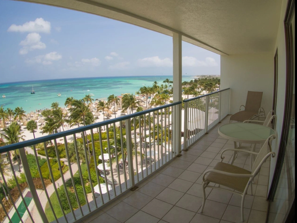 A tiled hotel balcony with outdoor seating offers a beautiful view of palm trees, white sand, and the clear ocean.