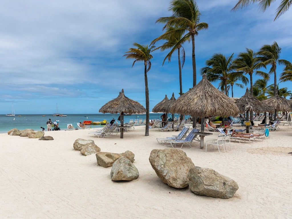 A relaxing beach scene featuring white sand, large rocks, thatched-roof umbrellas, and lounge chairs by the water.