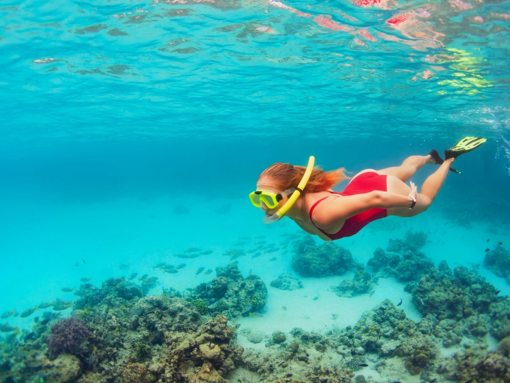 A woman wearing a red swimsuit and yellow snorkeling gear swims over a coral reef in clear blue water.
