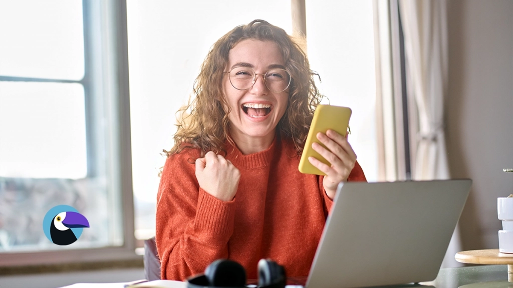 Excited woman holding a phone by a laptop, finding out why are timeshare resales so cheap online.