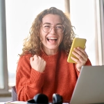Excited woman holding a phone by a laptop, finding out why are timeshare resales so cheap online.