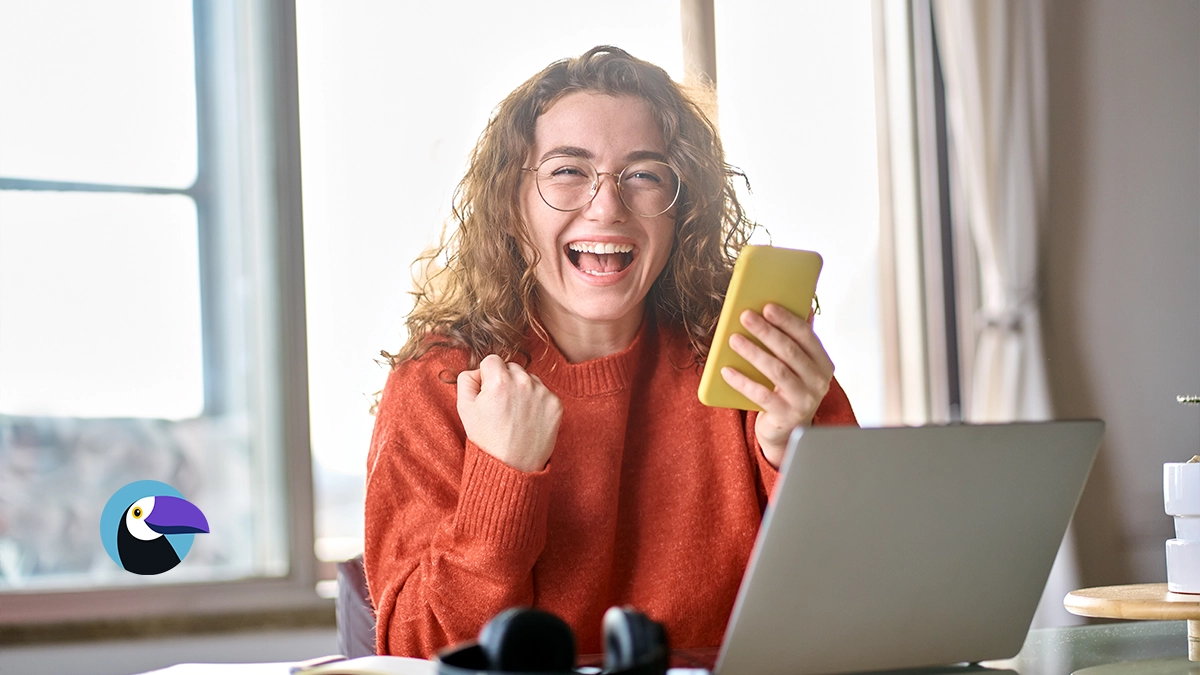 Excited woman holding a phone by a laptop, finding out why are timeshare resales so cheap online.