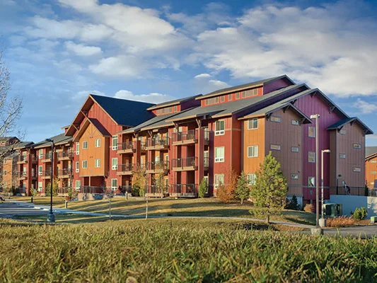 Multi-story apartment complex with red and purple siding, balconies, and pitched roofs surrounded by green lawn.