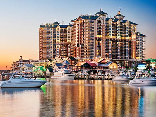 Waterfront marina at dusk with luxury high-rise hotel and boats reflected in calm water.
