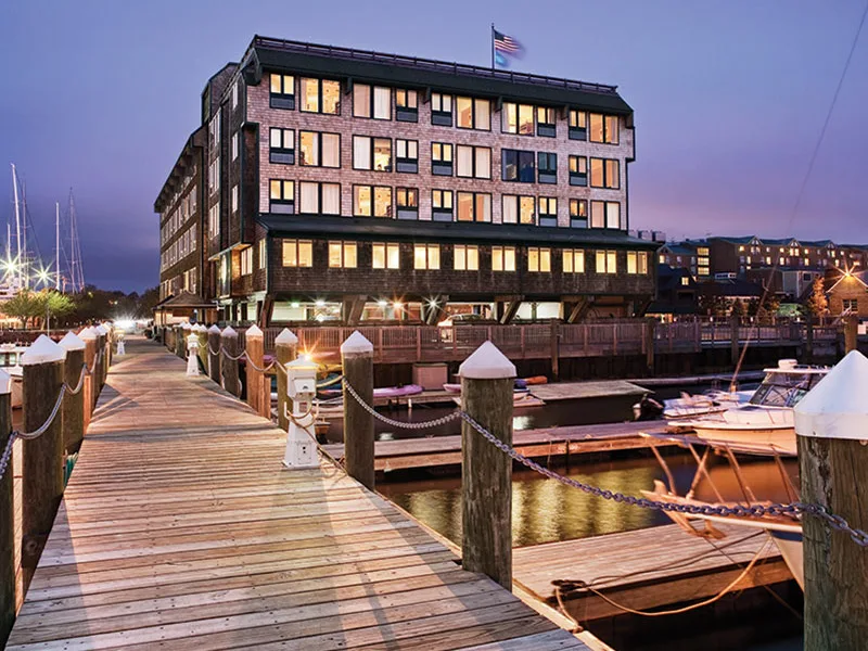 Wooden marina dock at dusk with boats moored beside a multi-story waterfront hotel with illuminated windows.
