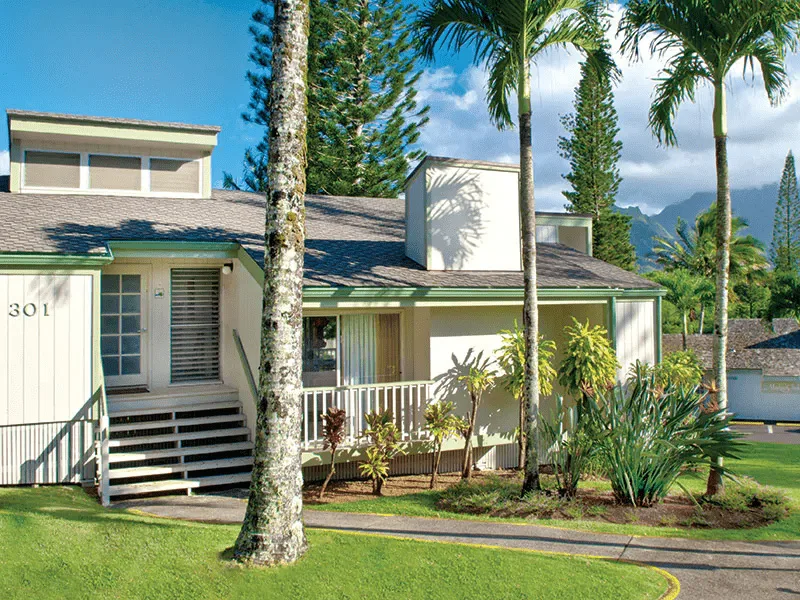 White two-story vacation resort building numbered 301 surrounded by palm trees and green lawn with mountain backdrop.